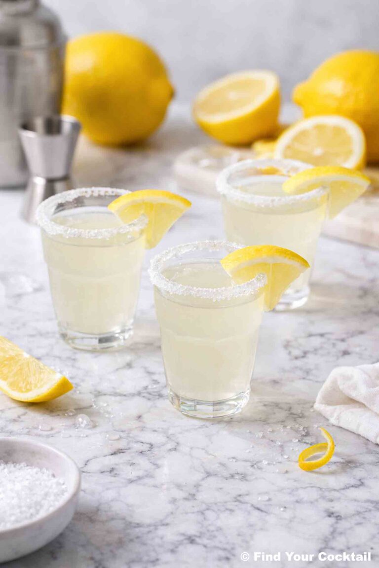 two shot glasses with sugared rims and a lemon wedge with lemons around and a cocktail shaker in the background.