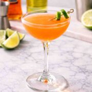 A naked & famous cocktail is a coupe glass with a lime twist and a bottle of aperol and yellow Chartreuse in the background.