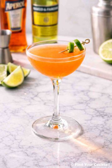 A naked & famous cocktail is a coupe glass with a lime twist and a bottle of aperol and yellow Chartreuse in the background.
