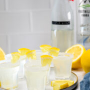 A plate of lemon cocktails in sugared rim glasses, garnished with lemon wedges, with bottles and fresh lemons in the background.