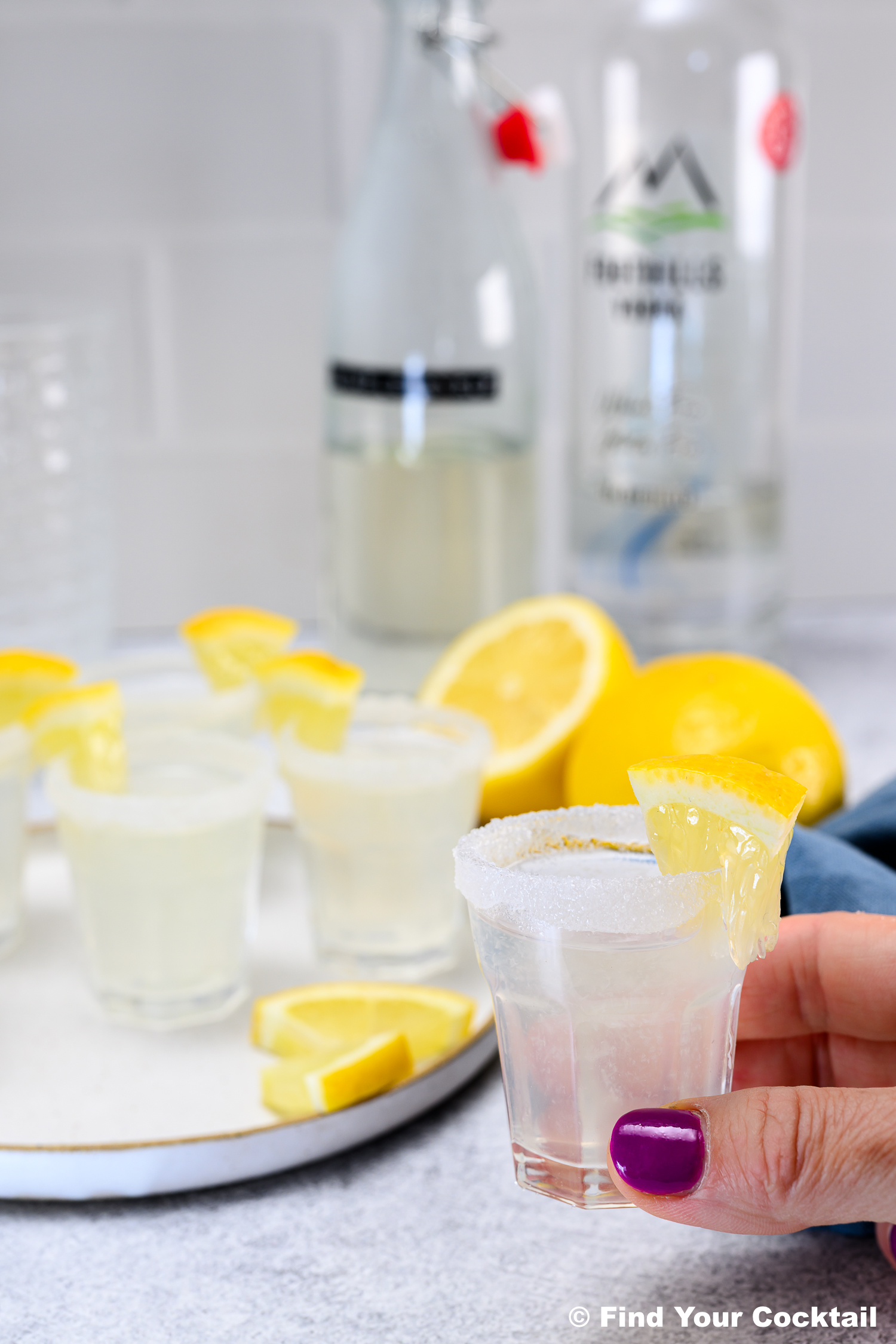 A hand with purple nail polish holds a sugar-rimmed shot glass with a lemon wedge; more similar shots and lemon slices are on a tray in the background.