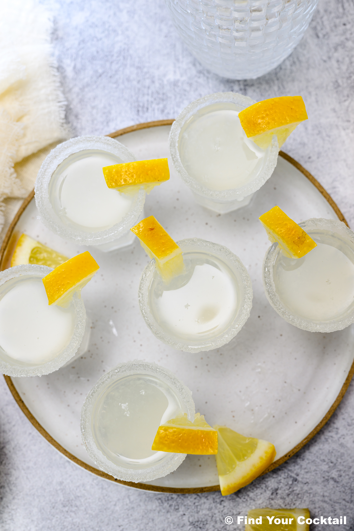 Six glasses with clear liquid and sugared rims, each garnished with a lemon wedge, arranged on a round white plate viewed from above.