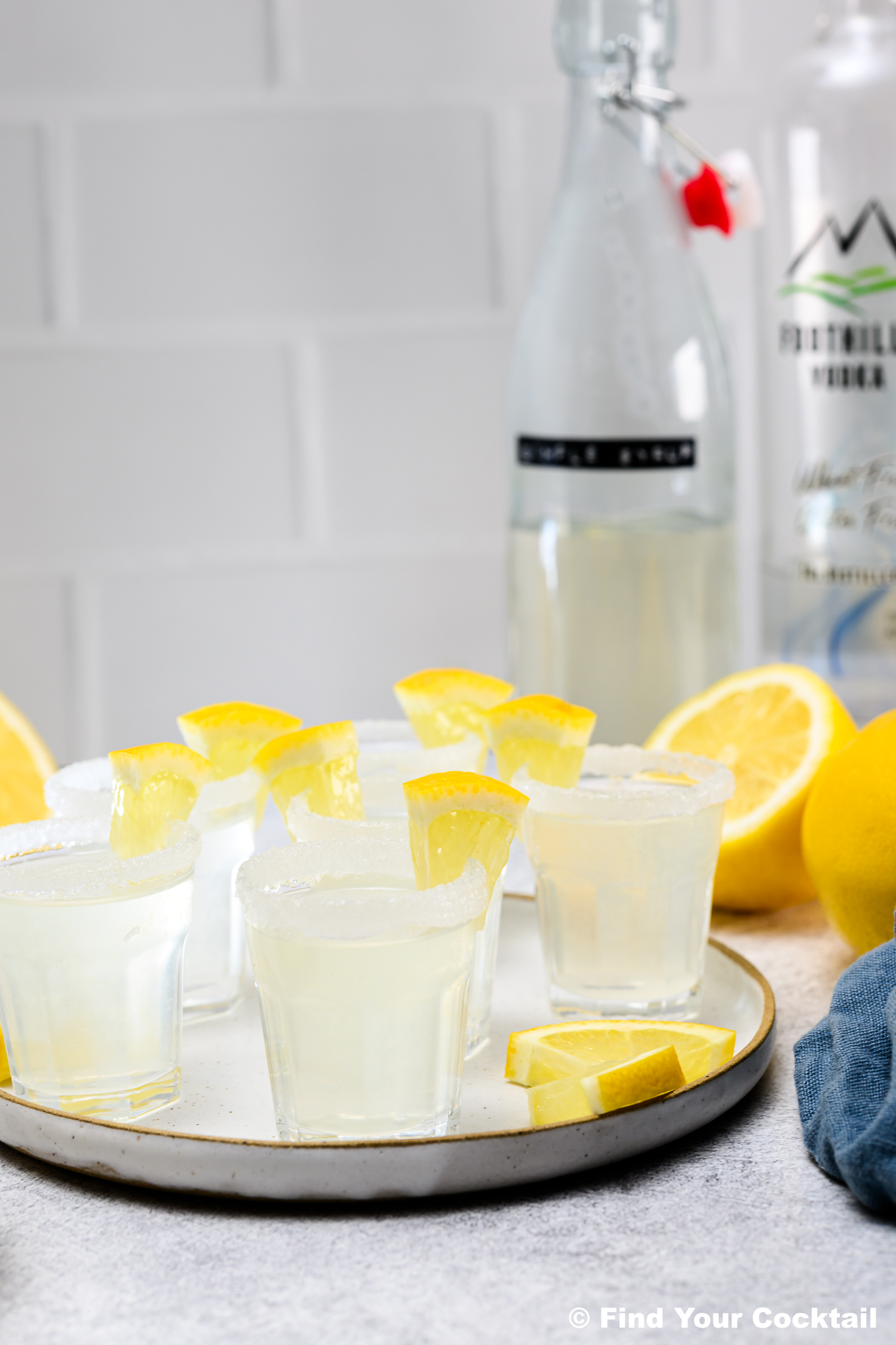 A plate of lemon cocktails in sugared rim glasses, garnished with lemon wedges, with bottles and fresh lemons in the background.