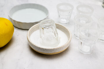 A shot glass is being dipped in sugar, next to other shot glasses with sugared rims, a dish of liquid, and a lemon on a white surface.