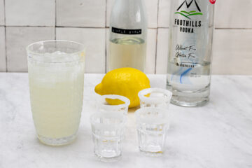 A glass of lemonade with ice, a lemon, a bottle of vodka, a bottle of simple syrup, and four sugar-rimmed shot glasses on a marble countertop.