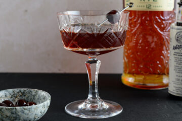 Recipe instruction photo: a manhattan cocktail in a coupe glass with a brandied cherry garnish and a bottle of rye on the side.