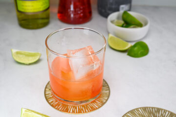 A glass with an orange-red cocktail and large ice cubes sits on a coaster, surrounded by lime wedges and bottles in the background.