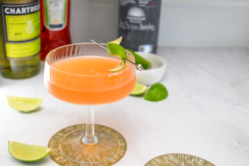 A cocktail in a coupe glass garnished with a lime wedge sits on a coaster, with liquor bottles and lime slices in the background.