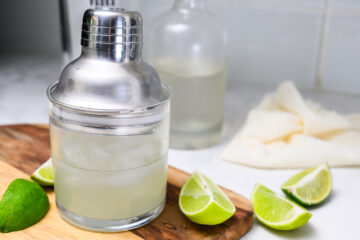 A cocktail shaker placed inside a glass filled with a clear drink and ice, surrounded by lime wedges on a wooden board. A bottle and cloth are in the background.