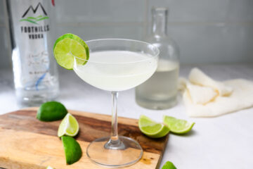 A cocktail glass with a clear drink garnished with a lime wheel sits on a wooden board, surrounded by lime wedges and vodka bottles in the background.
