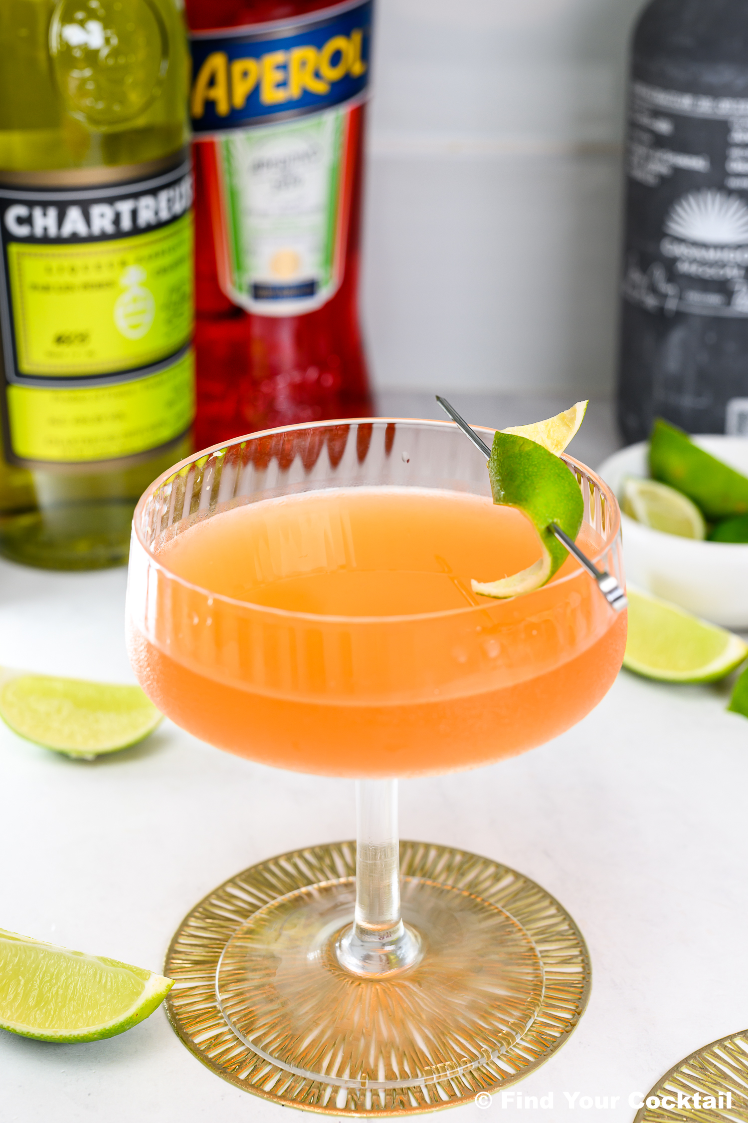 A coupe glass with an orange cocktail garnished with lime, surrounded by lime wedges and bottles of Chartreuse and Aperol in the background.