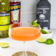 A coupe glass filled with an orange cocktail garnished with a lime twist, surrounded by liquor bottles and lime wedges on a white surface.