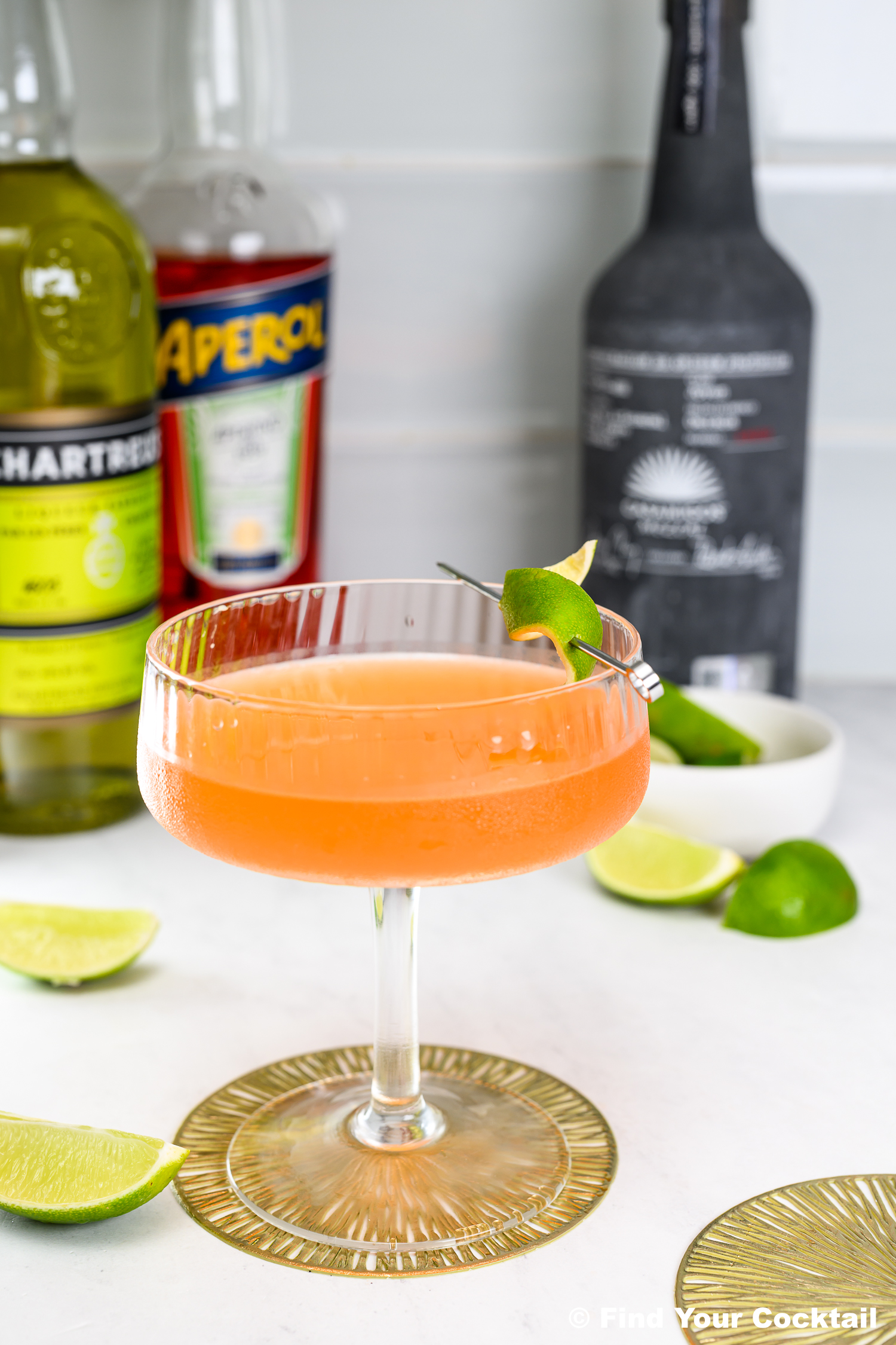 A coupe glass filled with an orange cocktail garnished with a lime twist, surrounded by liquor bottles and lime wedges on a white surface.