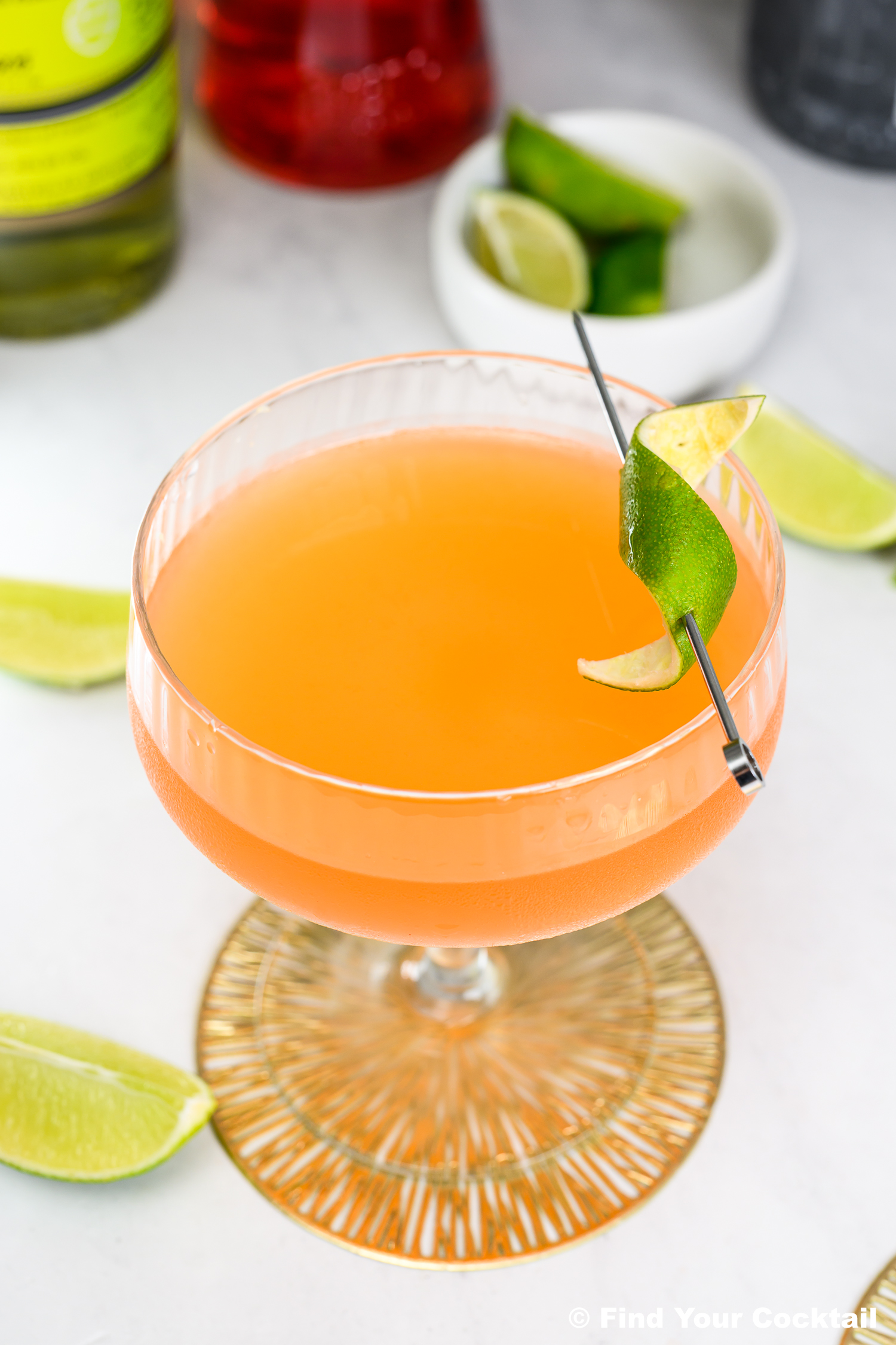 A coupe glass filled with an orange cocktail, garnished with a curled lime peel on a metal pick, sits on a white surface with lime wedges nearby.