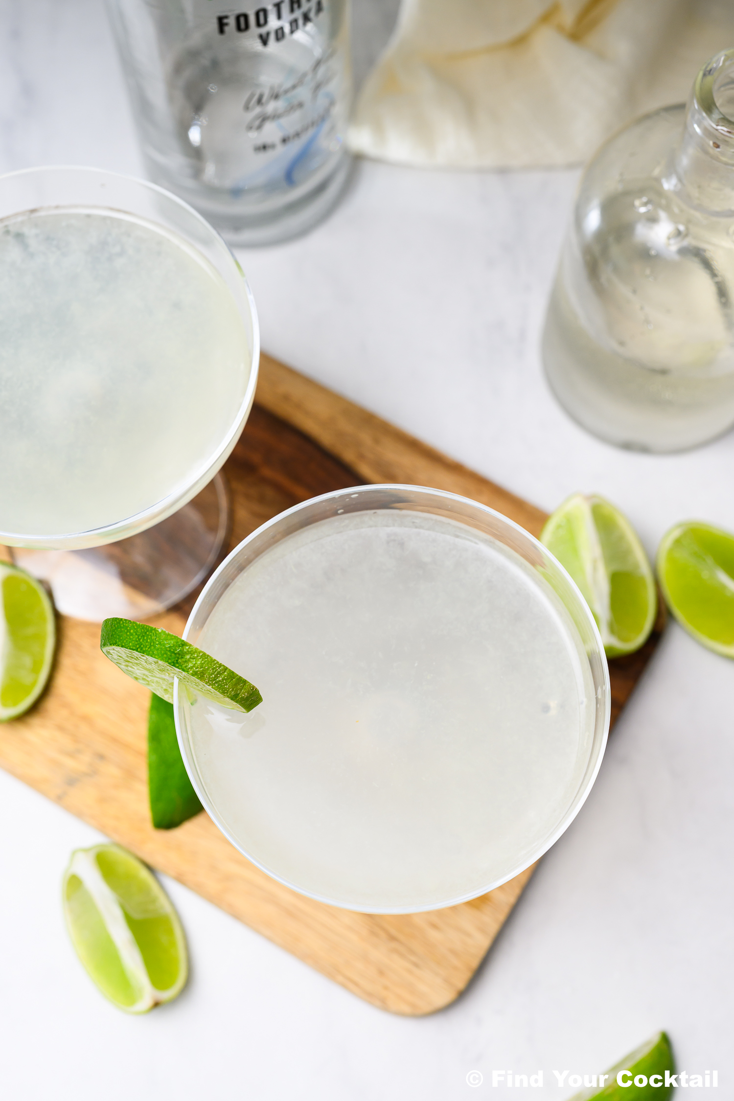 Two clear cocktails with lime wedges on a wooden board, surrounded by lime slices, a vodka bottle, and a clear bottle on a white surface.