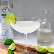 A clear cocktail in a coupe glass garnished with a lime wheel, surrounded by lime wedges, a bottle of vodka, and another glass bottle on a wooden board.