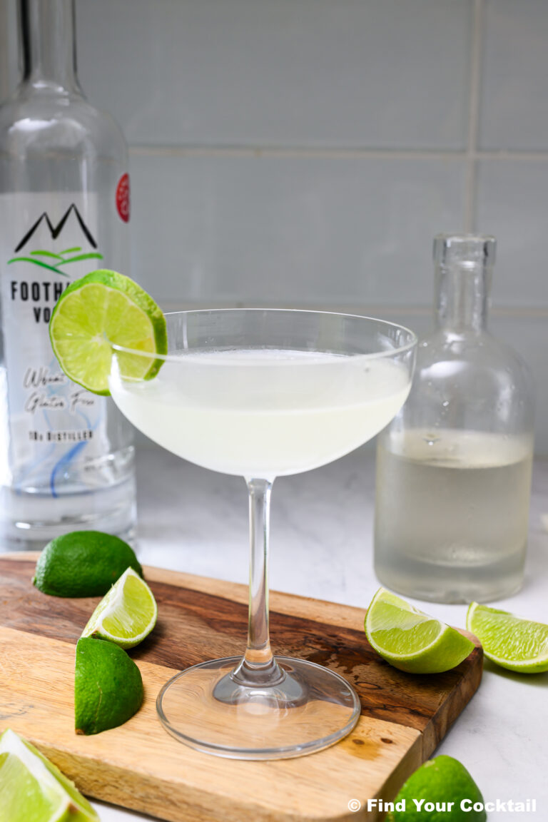 A clear cocktail in a coupe glass garnished with a lime wheel, surrounded by lime wedges, a bottle of vodka, and another glass bottle on a wooden board.