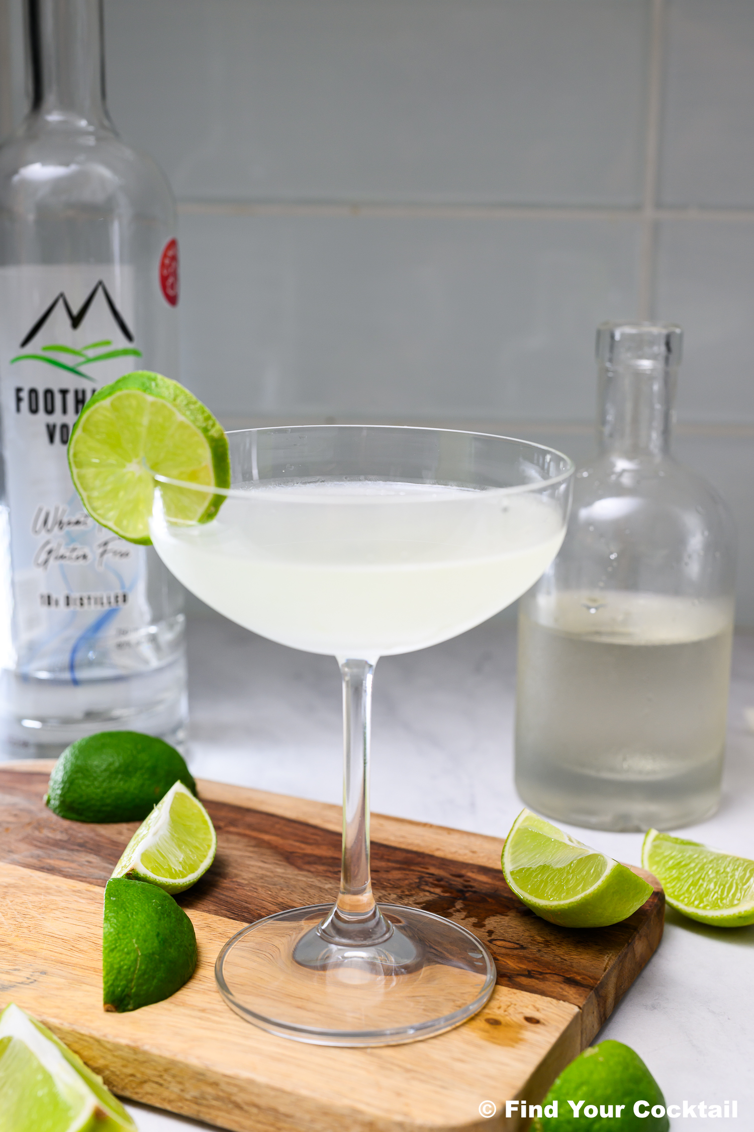 A clear cocktail in a coupe glass garnished with a lime wheel, surrounded by lime wedges, a bottle of vodka, and another glass bottle on a wooden board.