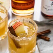 a cocktail in a rocks glass garnished with sliced apple and a cinnamon stick