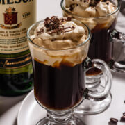 2 Irish coffees in tall glass mugs with a bottle of Irish whiskey in the background