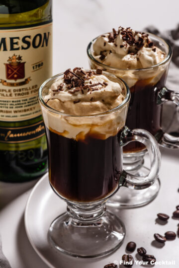 2 Irish coffees in tall glass mugs with a bottle of Irish whiskey in the background