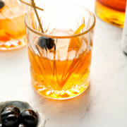 A glass of amber cocktail with a cherry garnish on a pick, next to a small dish of cherries and another drink in the background.