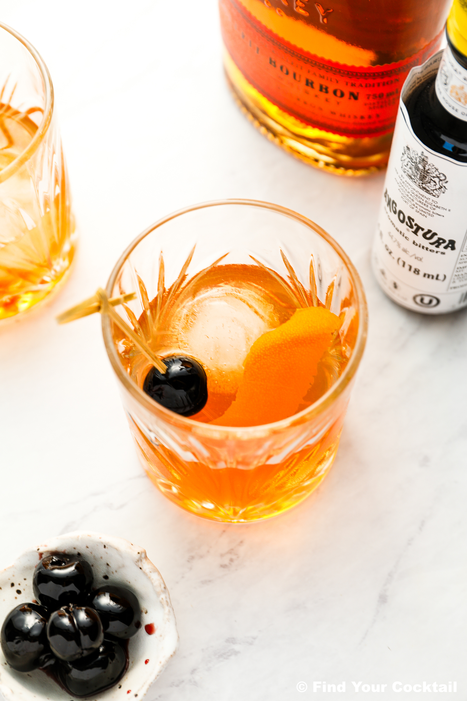 A glass of old fashioned cocktail with an ice sphere, orange peel, and cherry garnish, next to a bottle of bourbon, Angostura bitters, and a small bowl of cherries on a marble surface.