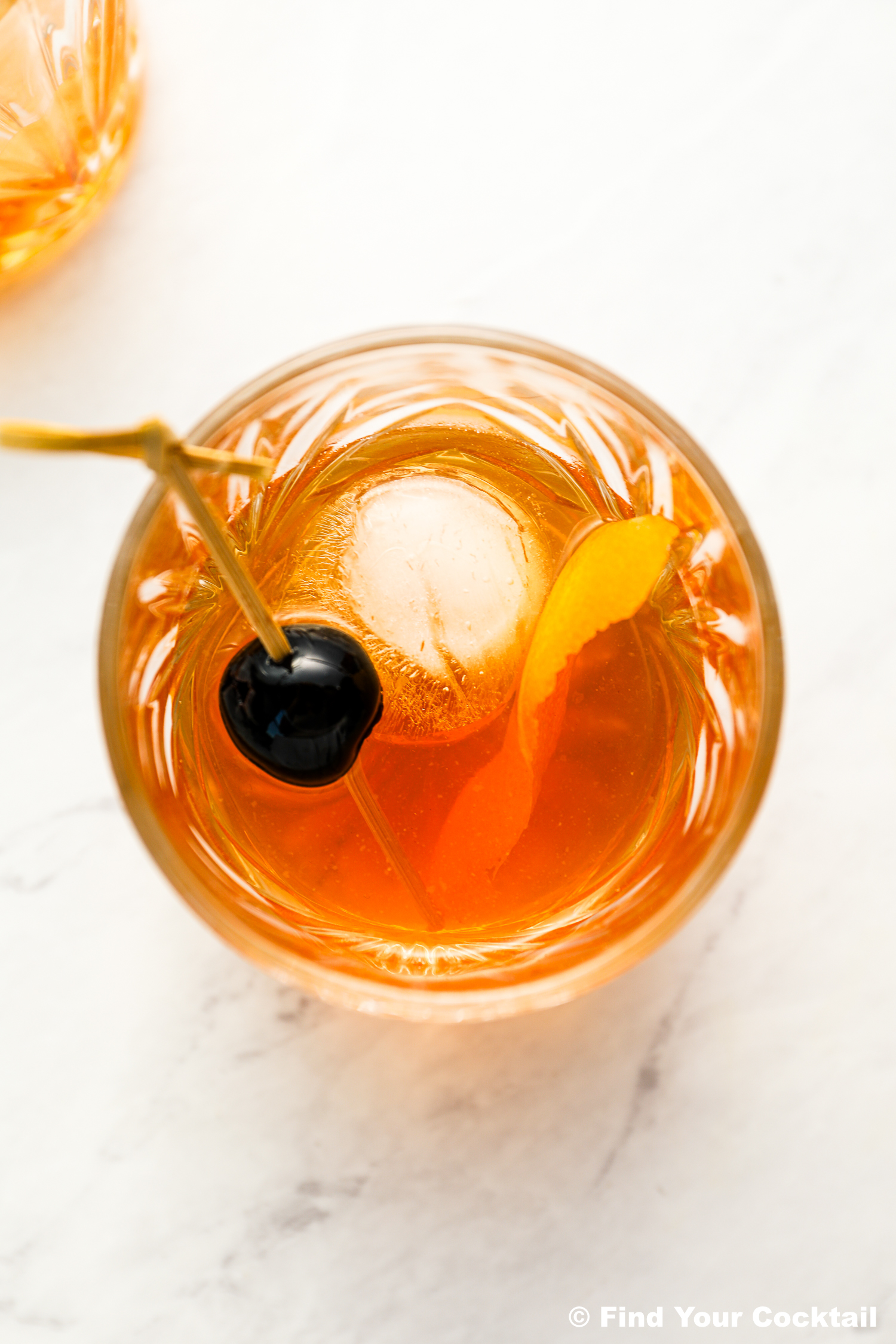A glass of amber-colored cocktail with a large ice sphere, garnished with an orange twist and a cherry on a cocktail pick, on a white surface.