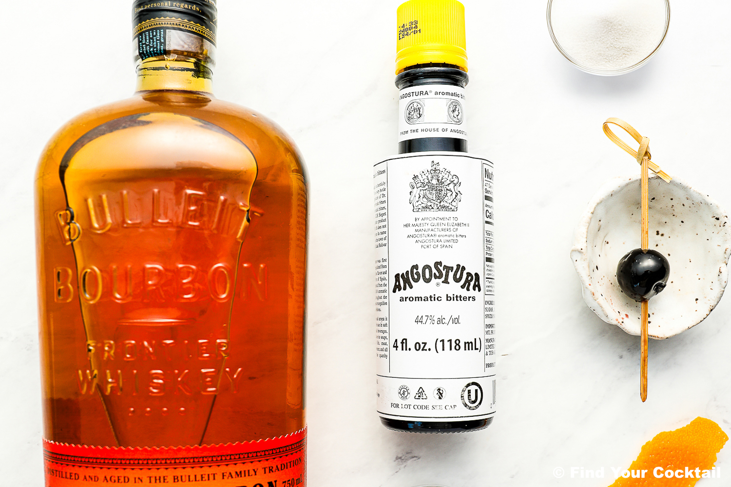 A bottle of Bulleit Bourbon, a bottle of Angostura aromatic bitters, a small bowl of sugar, and a cocktail cherry on a pick are arranged on a white surface.
