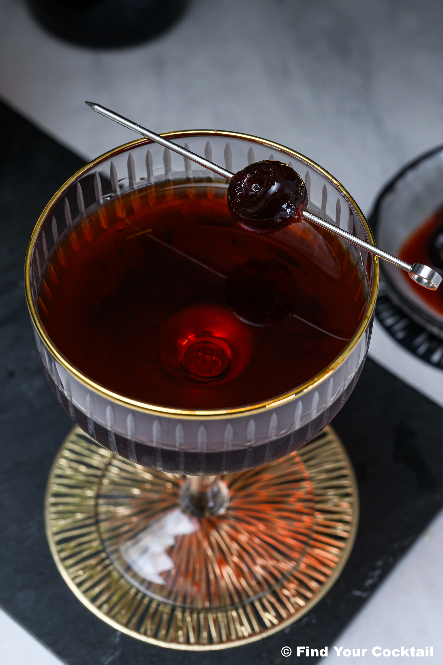 A coupe glass filled with a dark red cocktail, garnished with a single cherry on a metal pick, sits on a gold-rimmed coaster.