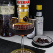A dark cocktail garnished with a cherry on a pick sits in a coupe glass, with bottles of rye, Averna, bitters, and a small dish of cherries in the background.