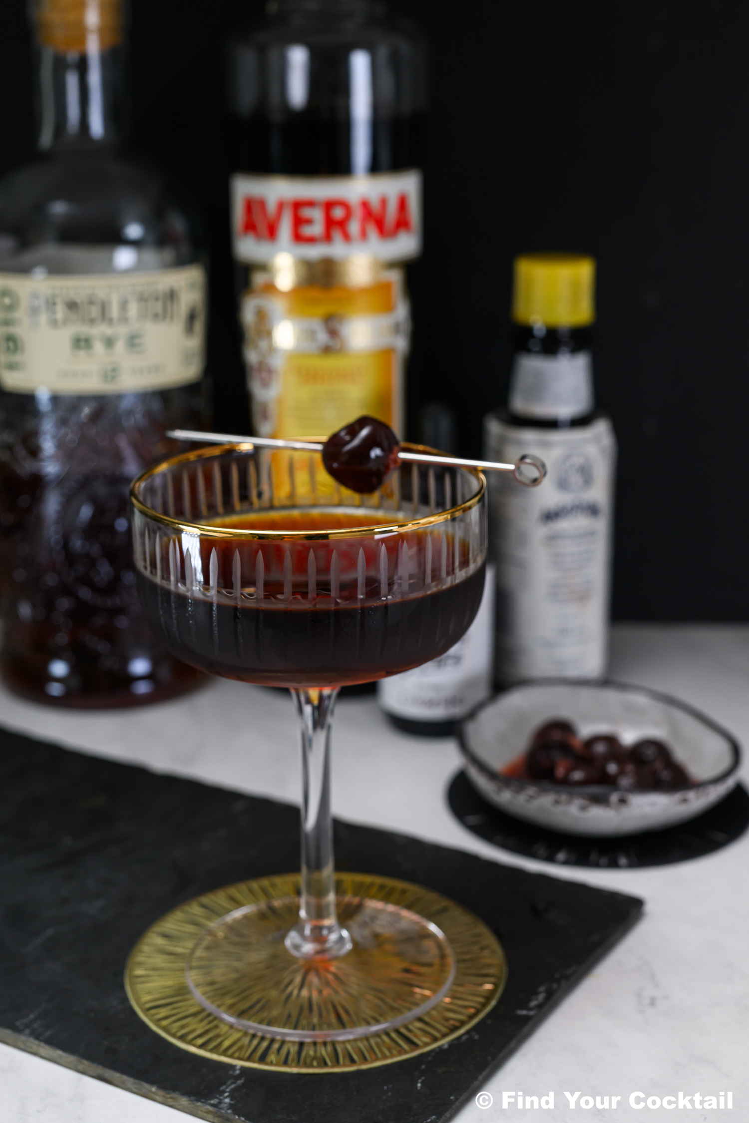 A dark cocktail garnished with a cherry on a pick sits in a coupe glass, with bottles of rye, Averna, bitters, and a small dish of cherries in the background.