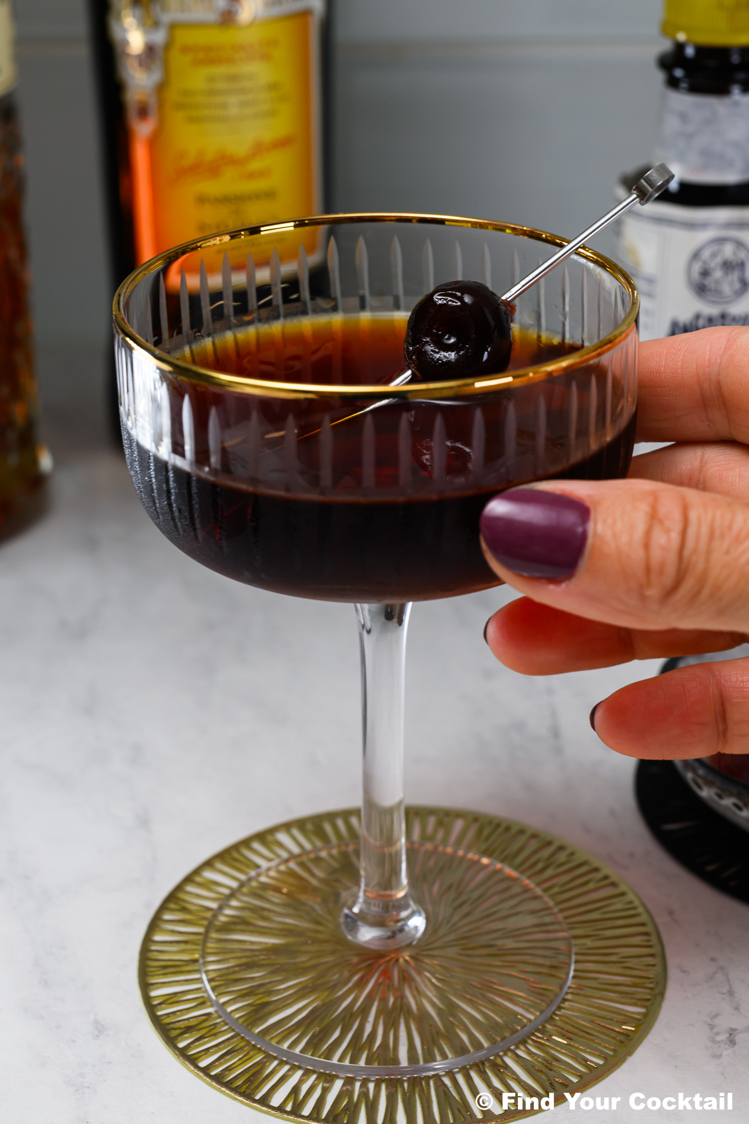 A hand holding a coupe glass filled with a dark cocktail, garnished with a cherry on a cocktail pick, set on a patterned coaster. Bottles are visible in the background.
