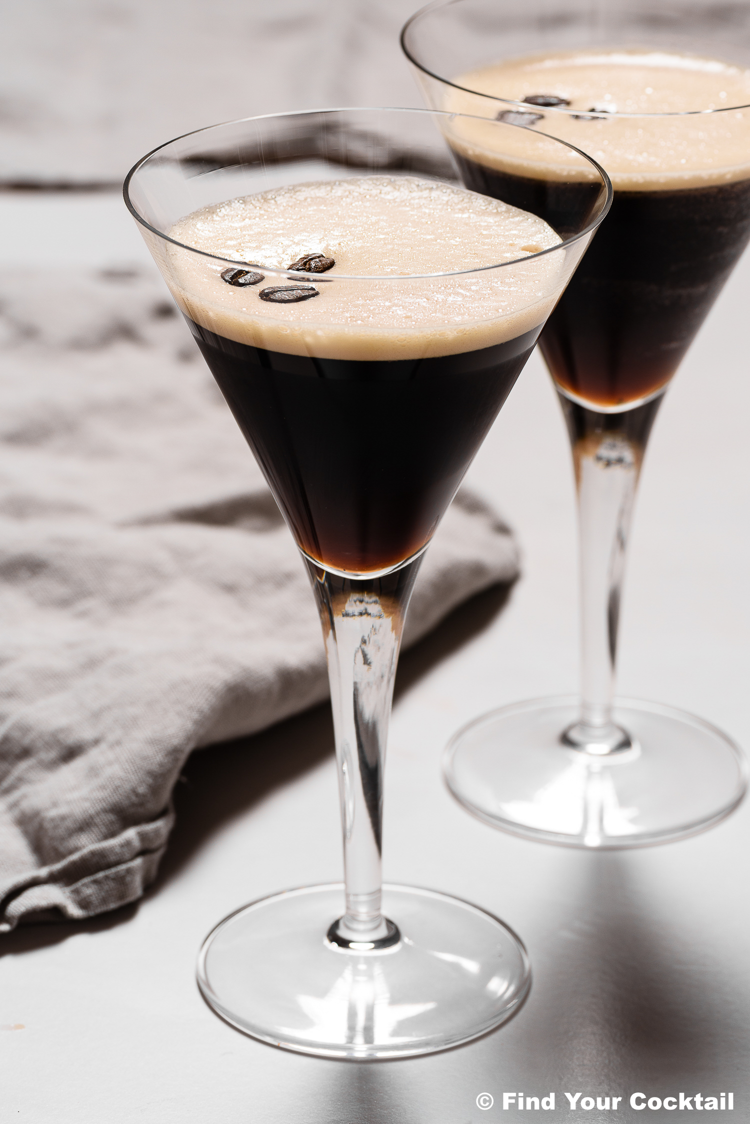 Two martini glasses filled with espresso martinis, topped with three coffee beans each, placed on a white surface next to a gray napkin.