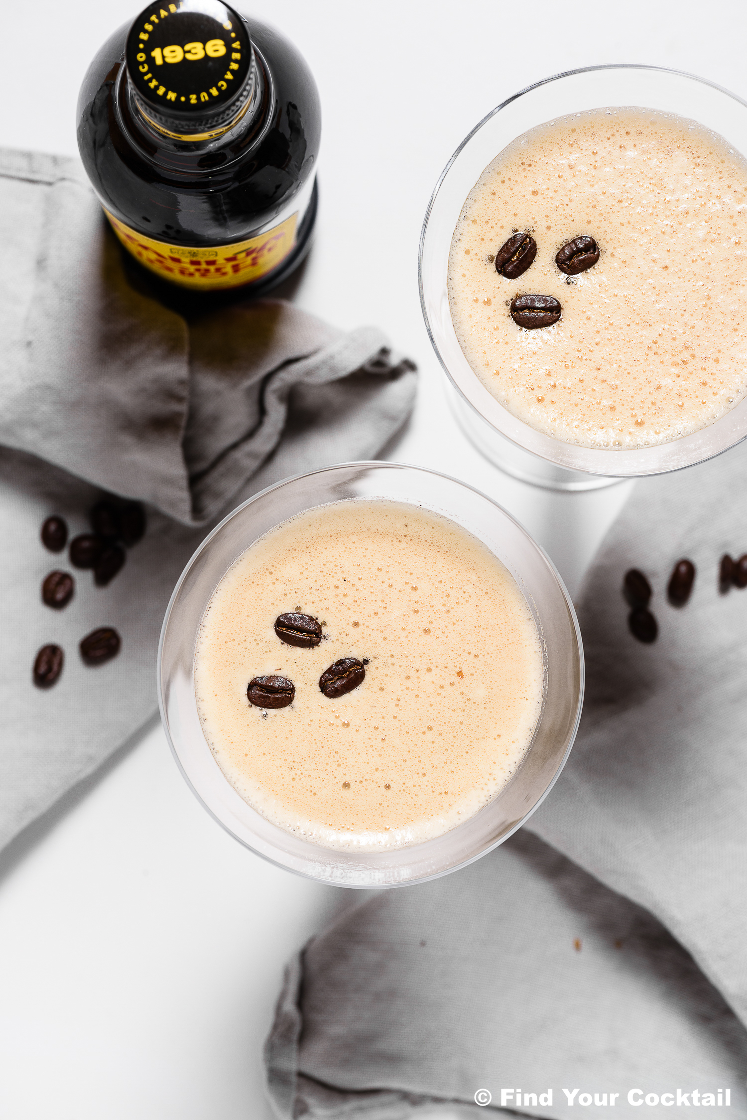 Overhead view of two frothy espresso martinis garnished with coffee beans, next to a bottle of liqueur and gray napkins on a white surface.