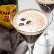 Two martini glasses filled with espresso martinis, each garnished with three coffee beans, sit next to a bottle of Kahl&uacute;a coffee liqueur on a white surface.