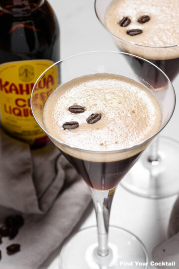 Two martini glasses filled with espresso martinis, each garnished with three coffee beans, sit next to a bottle of Kahlúa coffee liqueur on a white surface.