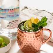 a copper mug filled with a gin mule and garnished with mint, lime, and ginger with a bottle of gin in the background