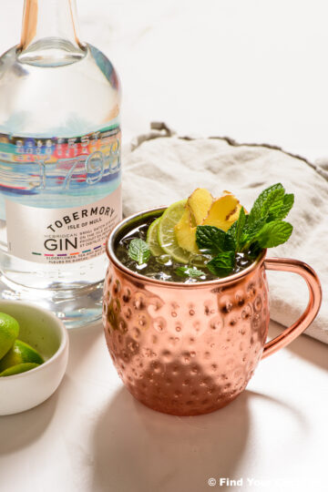 a copper mug filled with a gin mule and garnished with mint, lime, and ginger with a bottle of gin in the background
