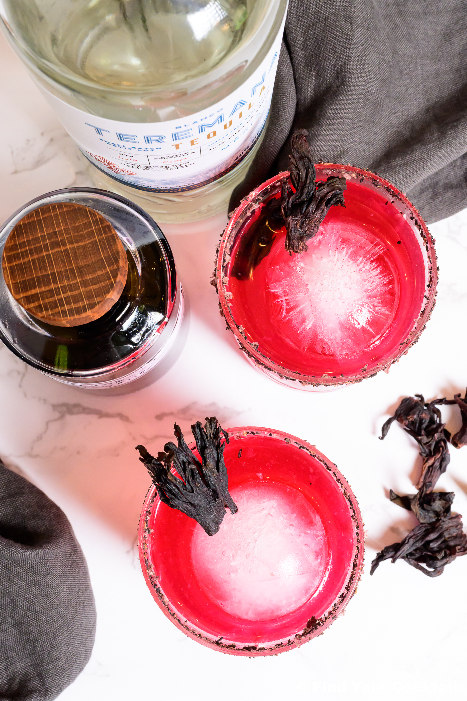 Looking down at 2 pink margaritas with large round ice cubes in rocks glasses and a bottle of tequila on the side.