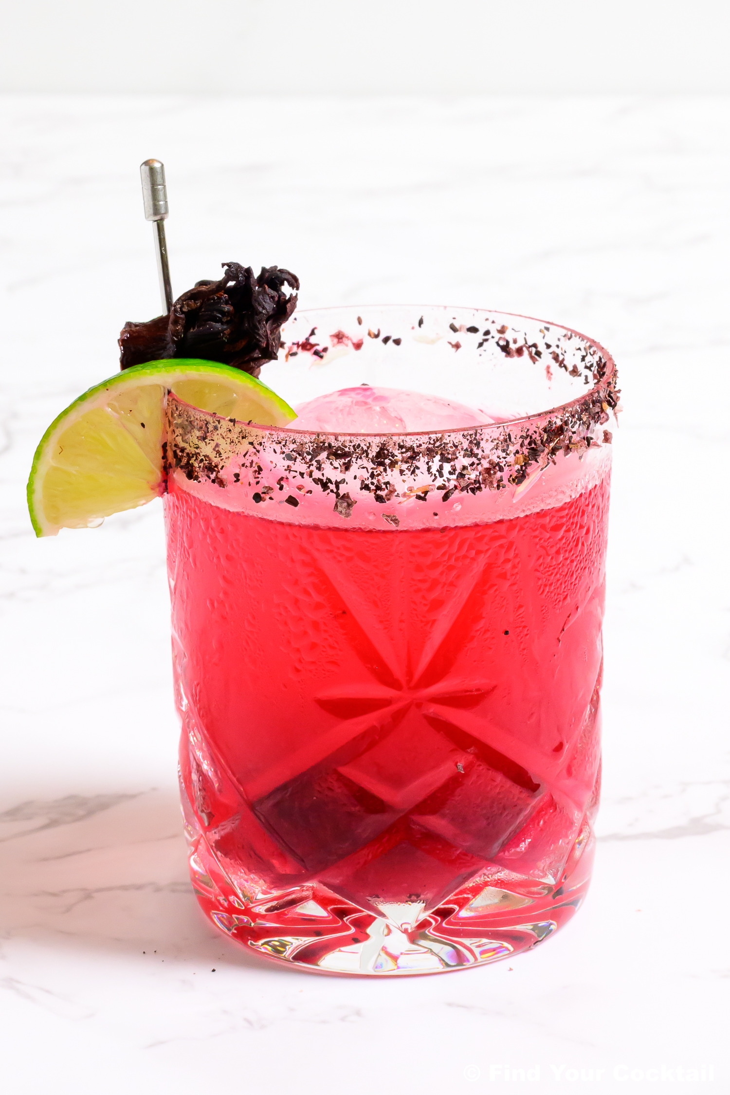 One bright fuchsia colored margarita in a rocks glass with a lime wedge and dried hibiscus flower on the side.