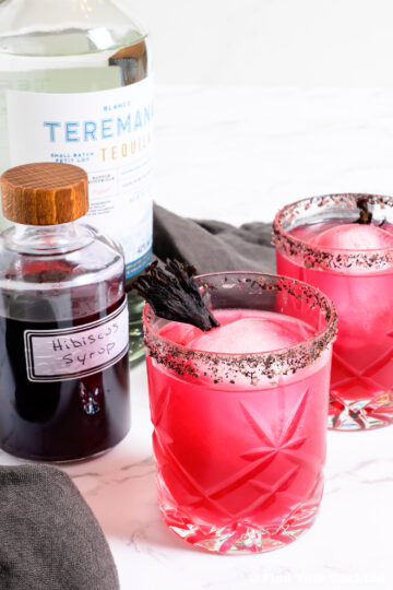 two hibiscus margaritas next to a bottle of hibiscus syrup and a bottle of tequila