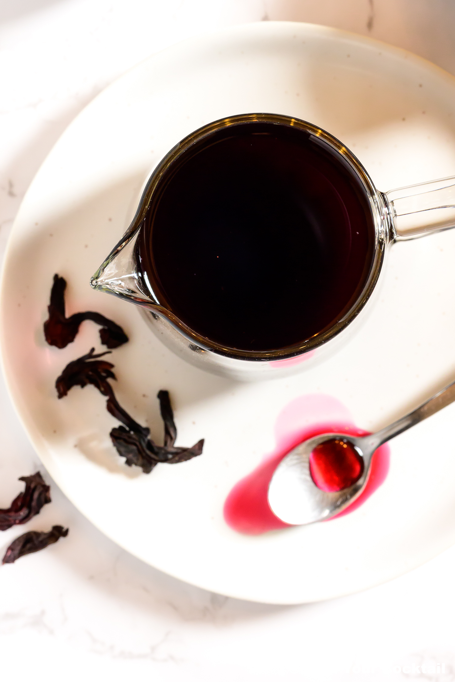 looking down on a small pitcher of hibiscus syrup on a white plate with a spoon of the syrup on the side.