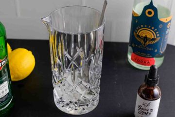 Recipe instruction photo: a cocktail mixer surrounded by a vodka bottle, dry vermouth, and orange bitters.