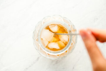 A hand stirs a glass of iced tea with ice cubes using a metal spoon, viewed from above on a white surface.