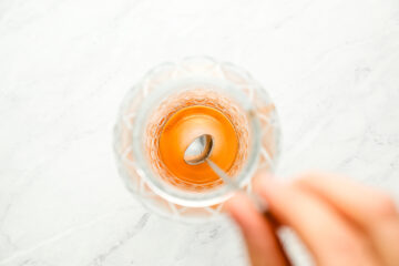 A hand stirs an amber-colored liquid in a faceted glass with a metal spoon, viewed from above on a light marble surface.