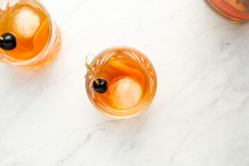 Top-down view of two glasses filled with an orange cocktail, ice, an orange peel, and a dark cherry garnish on a light marble surface.