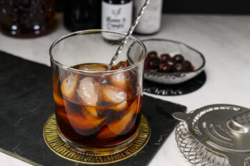 A glass of dark cocktail with ice and a stirring rod on a coaster, with a cocktail strainer and a bowl of cherries in the background.