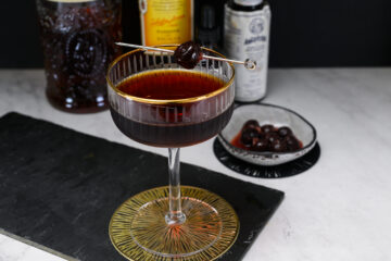 A classic cocktail in a coupe glass, garnished with a cherry on a cocktail pick, sits on a black slate with cocktail ingredients and a dish of cherries in the background.