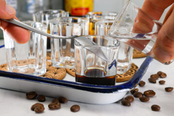 A person pours clear liquid over a spoon into a glass containing dark liquid, with other empty glasses and coffee beans on a tray nearby.